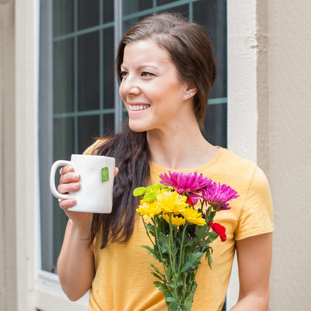 A woman in a yellow shirt stands by a window, smiling and holding a white mug with a tea bag. She holds a colorful bouquet of flowers in her other hand.